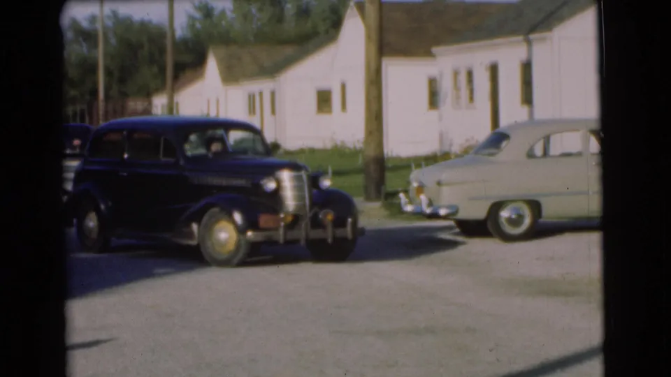 A black mid-century sedan crosses the foreground in 1951 Chatham, with a pale car and white houses behind it.