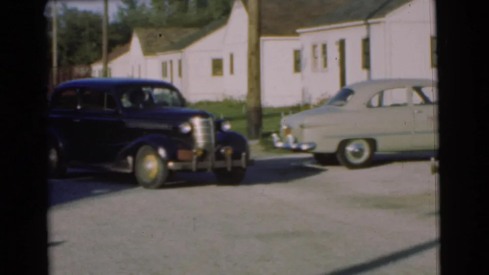 The black sedan exits toward the left edge of the Chatham parking-lot view, with the pale car and clapboard houses still visible.