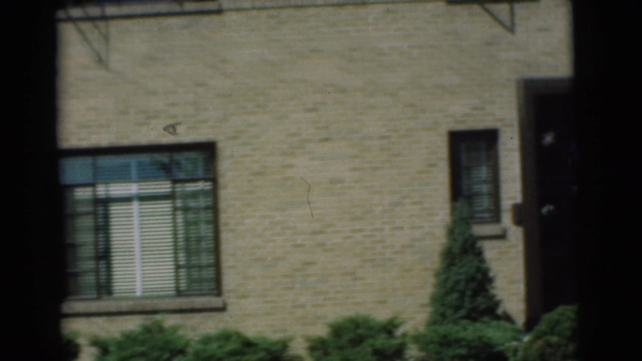 1951 Chatham, New Jersey home-movie frame showing a brick house wall, venetian-blind window, shrubs, and part of the front entrance.