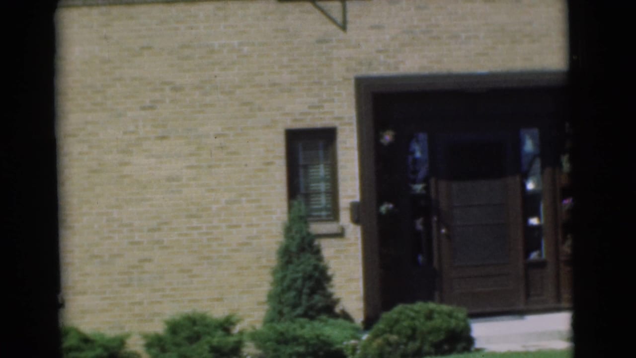 Brick house front entrance with dark door, narrow window, shrubs, and evergreen.