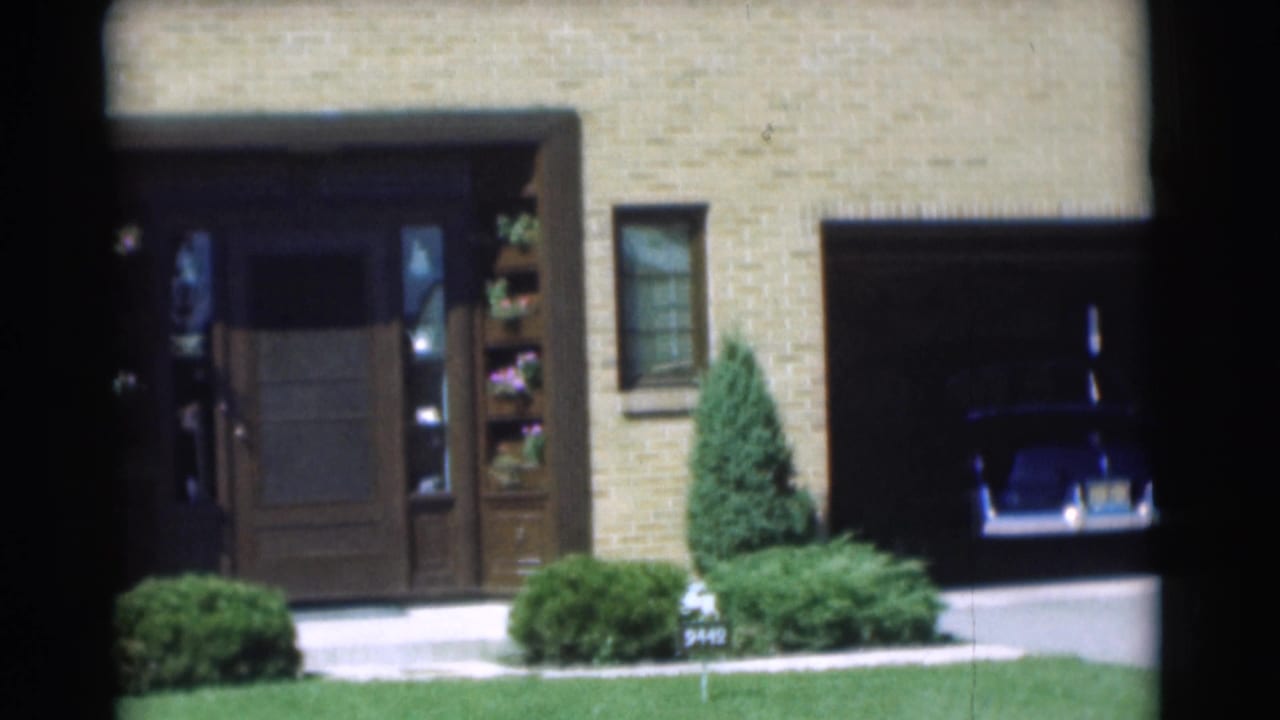 1951 Chatham, New Jersey home-movie frame showing a brick house, open garage, shrubs, lawn, and a dark sedan inside.
