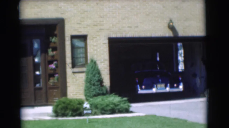 A mid-century dark sedan starts backing out of the garage at a brick residence in Chatham, New Jersey.