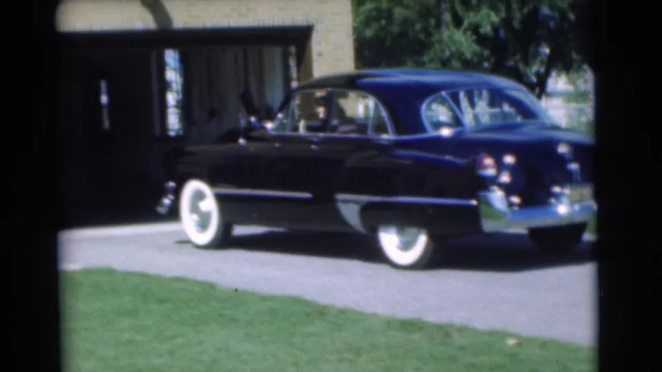 The dark 1950s sedan backs down the driveway, with a person faintly visible inside and the brick house behind it.