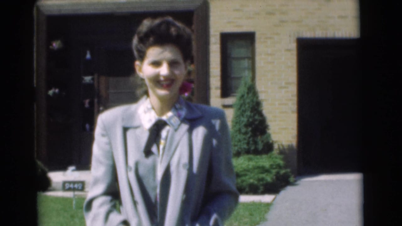 In Chatham, New Jersey in 1951, a woman in a light suit smiles toward the camera outside a brick suburban house.