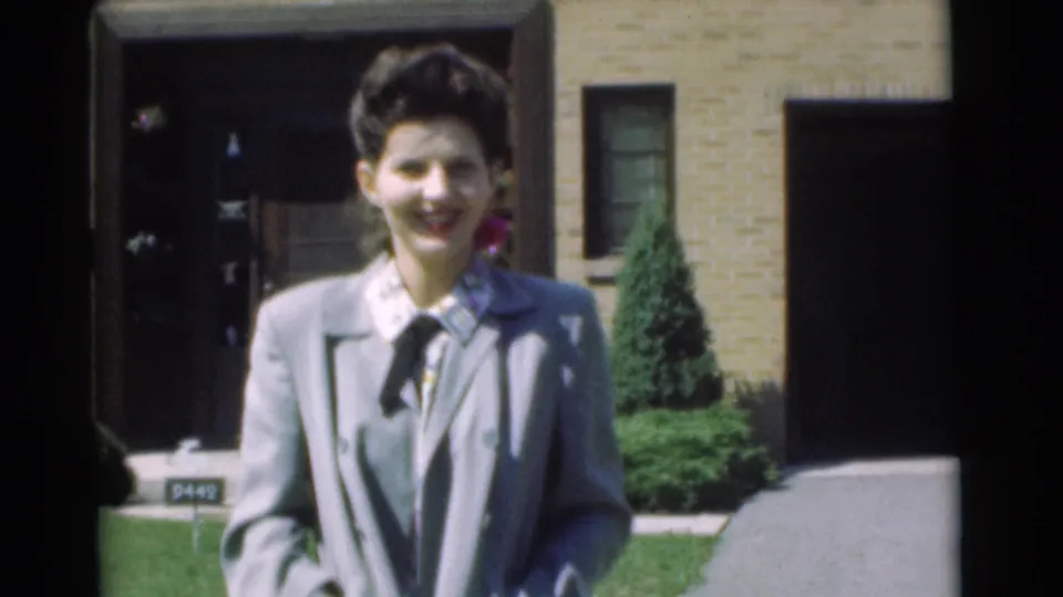 In Chatham, New Jersey in 1951, a woman in a light suit smiles toward the camera outside a brick suburban house.