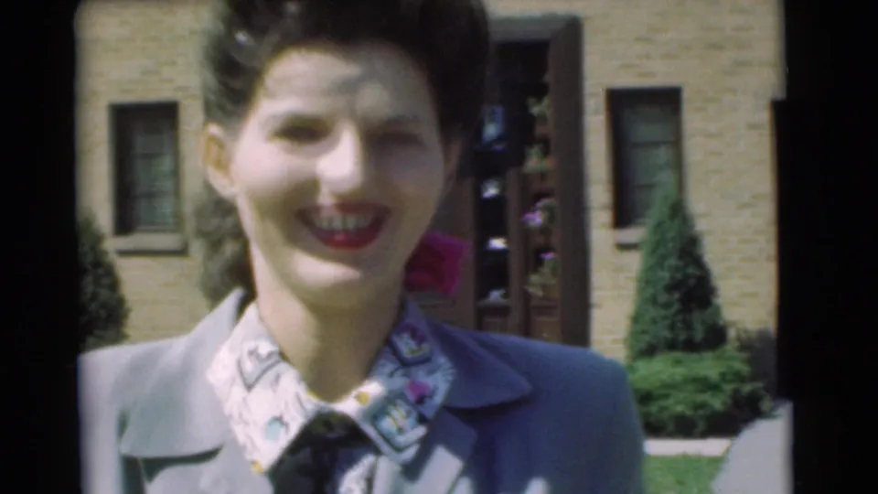 The 1951 home-movie frame moves closer to the smiling woman, showing her styled hair, red lipstick, and patterned blouse outside the house.