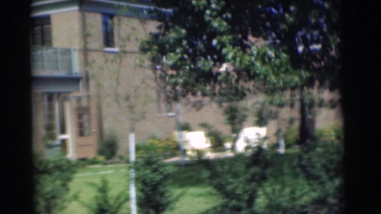 1951 Chatham, New Jersey 8mm frame showing a brick building with balcony, green lawn, shrubs, trees, and white outdoor chairs.