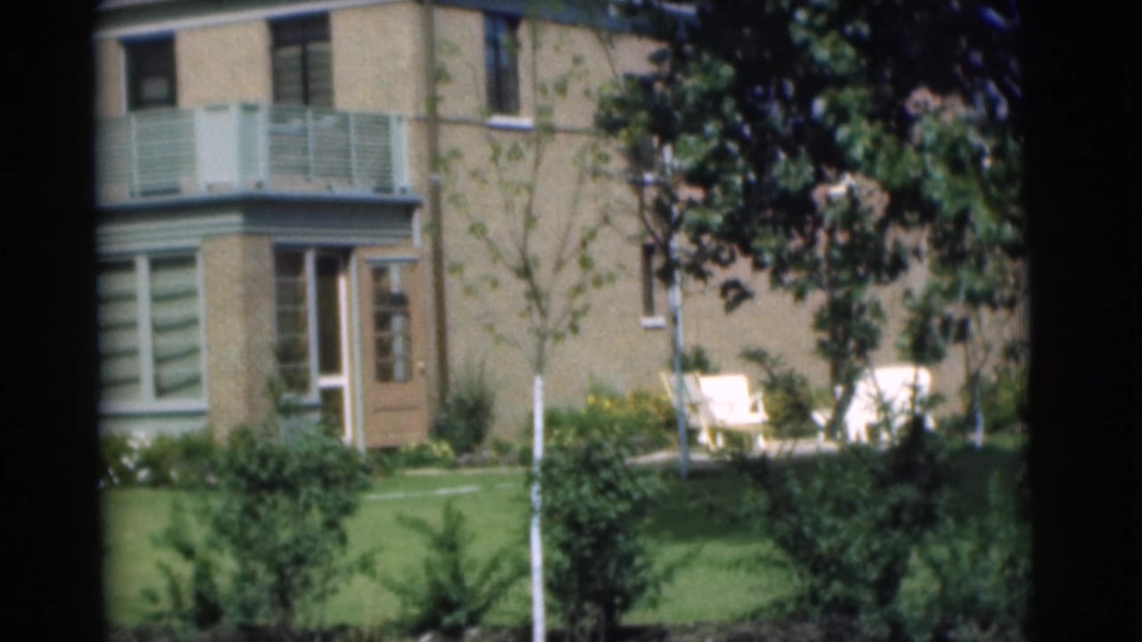 Sunny brick building exterior with pale balcony railing, doorway, lawn, shrubs, trees, and two white chairs.