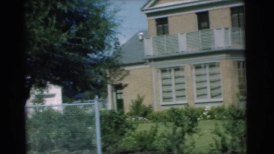 1951 Chatham, New Jersey frame showing the side of a brick building with balcony railing, shuttered windows, lawn plantings, shade, and fence.