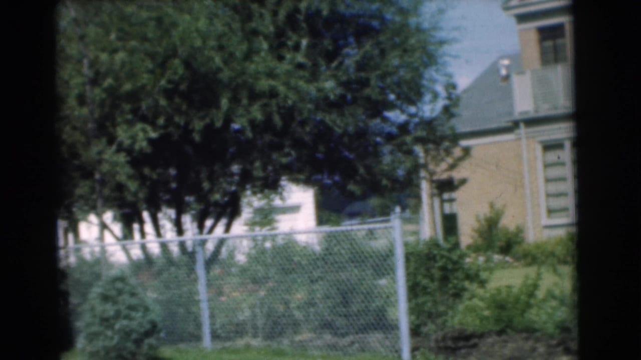 1951 Chatham, New Jersey home-movie frame showing trees, a chain-link fence, shrubs, and part of a tan residential house.