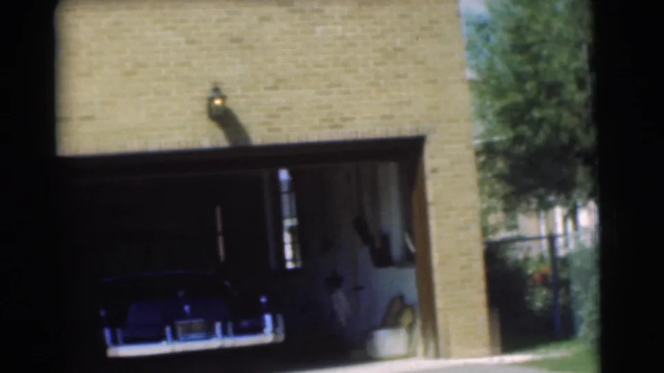 1951 Chatham, New Jersey home-movie frame showing a dark car parked inside an open garage of a tan brick house.
