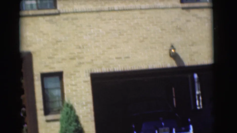 1951 Chatham, New Jersey frame showing the tan brick house facade, open garage, dark parked car, window, and shrub.