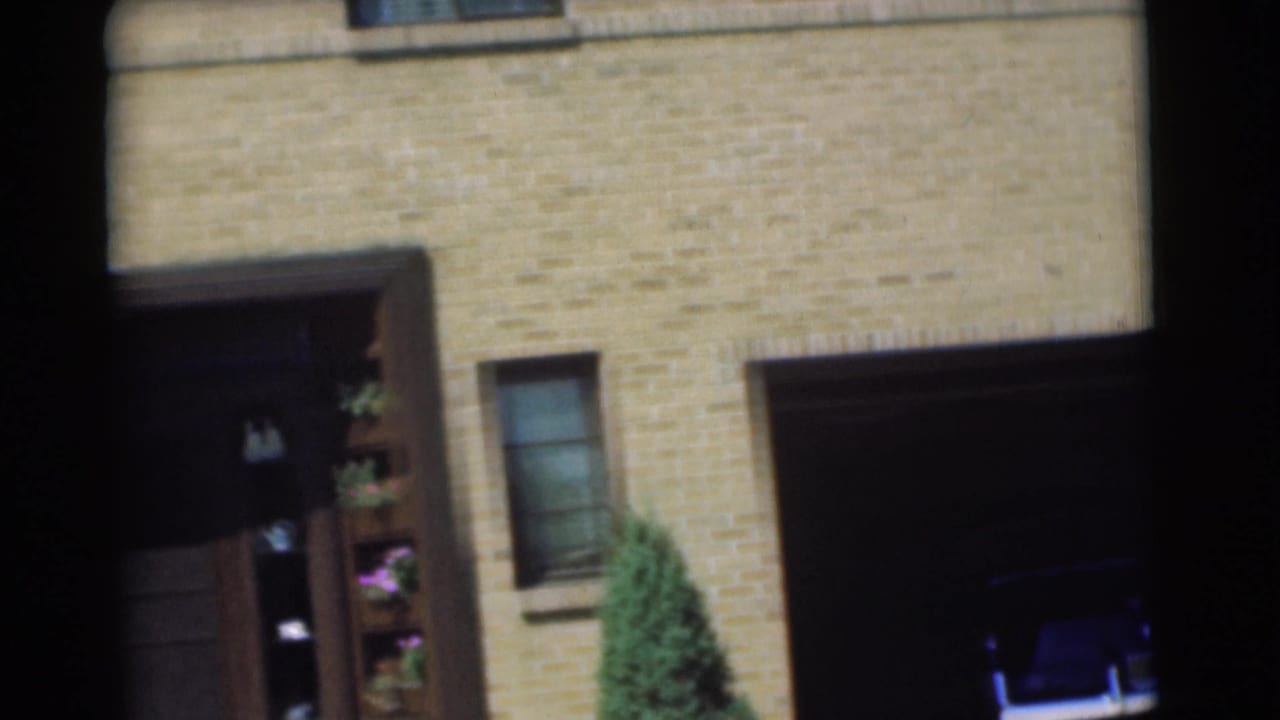 Brick house facade with dark recessed doorway, small window, shrubs, and garage opening at the right.