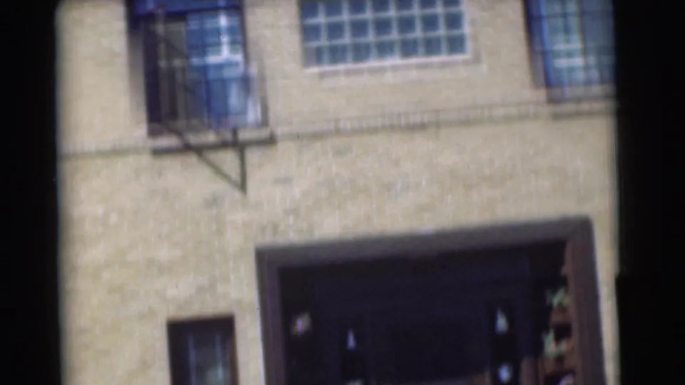 Handheld 1951 Chatham home-movie frame tilted across the brick house facade, showing a glass-block window, doorway edge, and lower window.