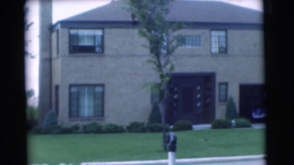 Wide 1951 Chatham, New Jersey view of a two-story brick suburban house with lawn, young tree, trimmed shrubs, recessed entry, and side garage.