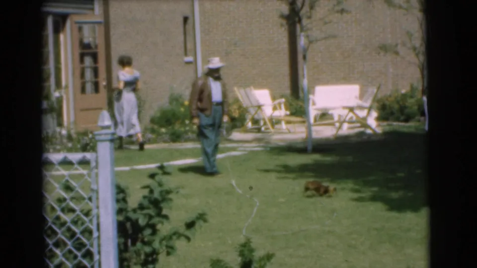 1951 Chatham, New Jersey backyard view with a hatted man near a brick house, a woman by the door, and a puppy on the grass.