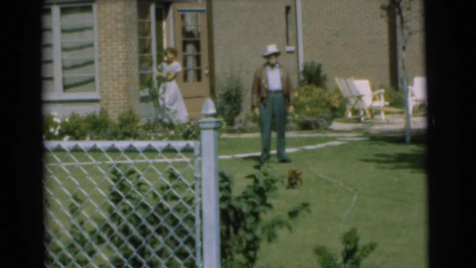 1951 Chatham, New Jersey backyard frame with a man in a hat, a woman at the house, and a small brown puppy on the lawn.