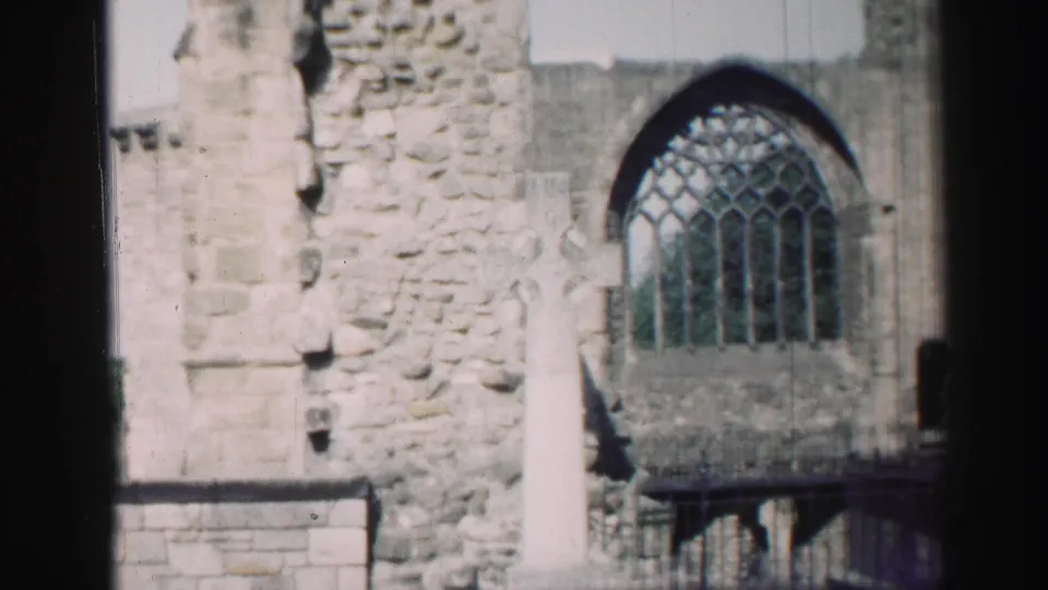 Soft 1950s 8mm frame of the church exterior, with the cross centered near rough masonry and an arched stained-glass window.