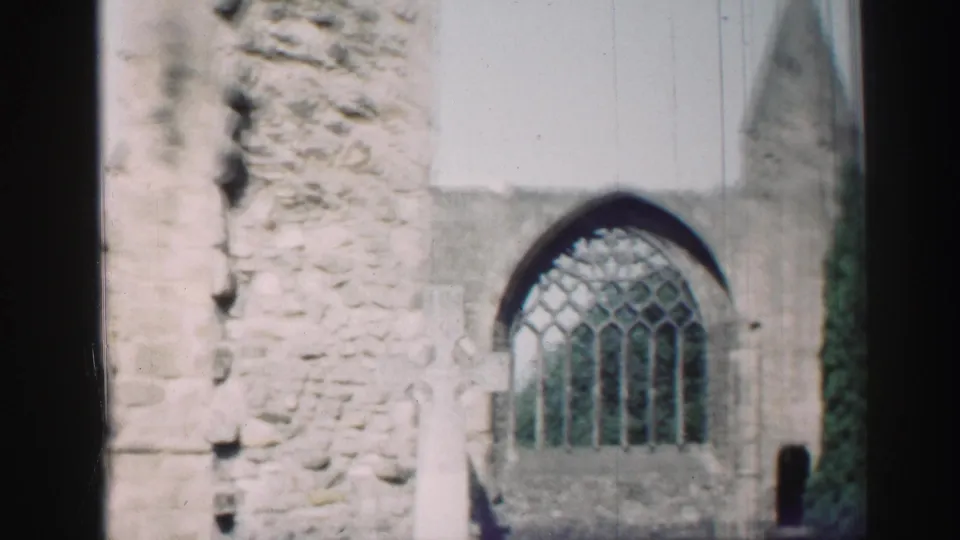1952 Scotland home-movie frame showing weathered church ruins, a Gothic arched window, pale grave markers, and dark film-edge borders.