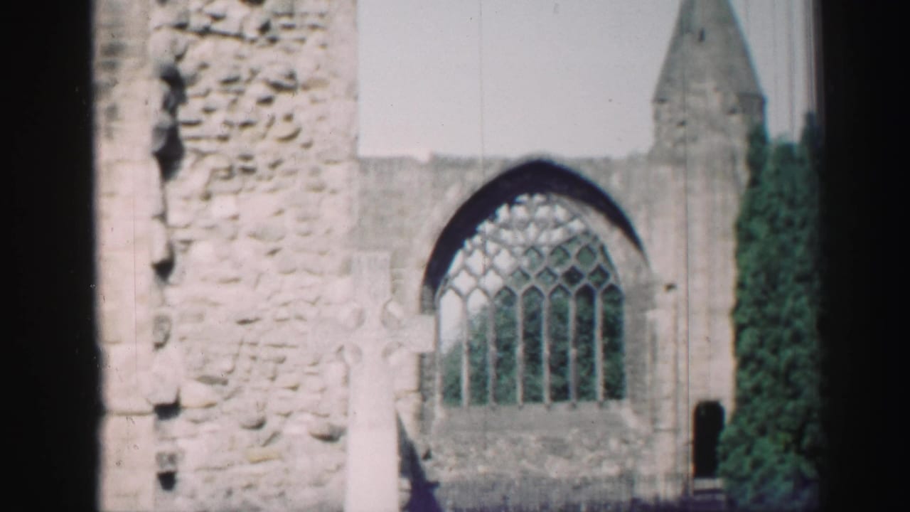 1952 Scotland frame of church ruins with Gothic window tracery, a cross-shaped grave marker, dense greenery, and a pointed roofline in the background.