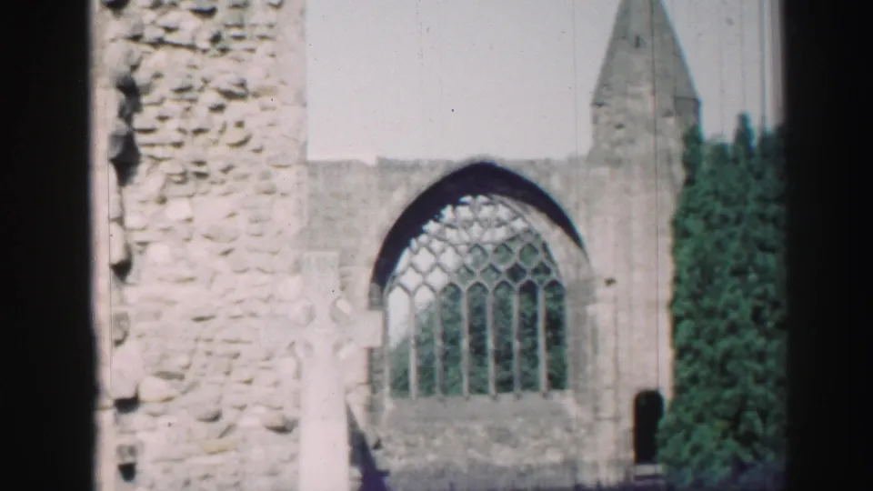 1952 Scotland home-movie still showing rough church masonry beside an arched window, a pale grave marker, and tall evergreen foliage.