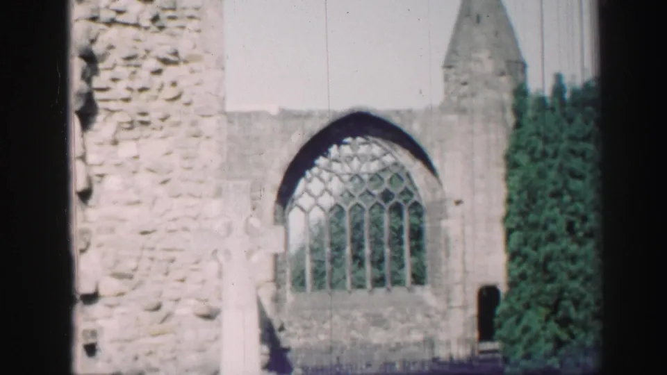 Final 1952 Scotland frame holding on the ruined church wall, Gothic arched window, pale grave marker, dark doorway-like gap, and dense greenery.