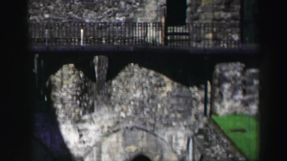 1952 8mm Edinburgh frame showing a higher view of ruined stone walls below a metal railing, with grass and dark film shadow at the edge.