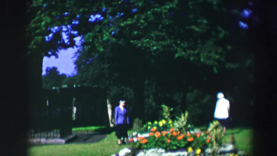 1952 Scotland garden scene with two distant visitors walking near a lawn, trees, bright flowerbeds, and historic stone ruins.
