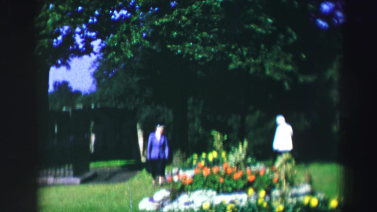 Two women walk near a bright flowerbed in a shaded garden lawn.