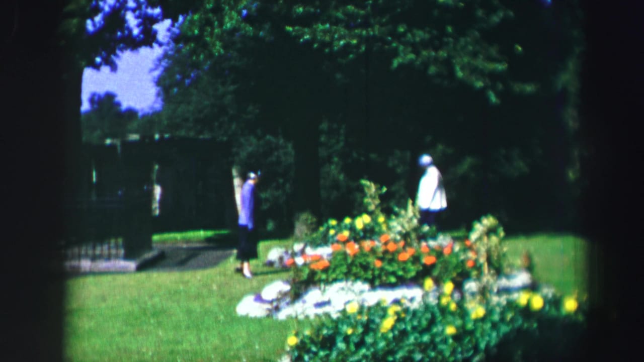 Two women stand apart beside a colorful flowerbed and lawn.