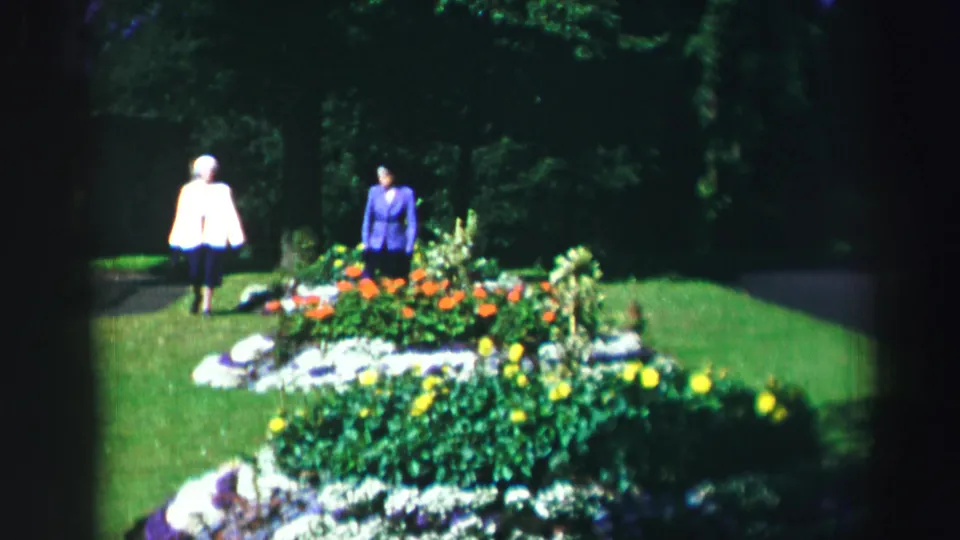 Two adult women in mid-century dress stand by a bright flowerbed on a lawn, framed by trees and deep shade.