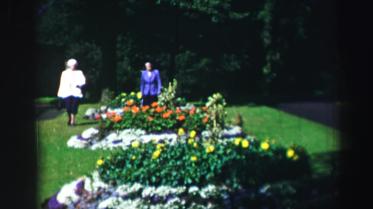 Two women stand near a colorful flowerbed on a sunny lawn.