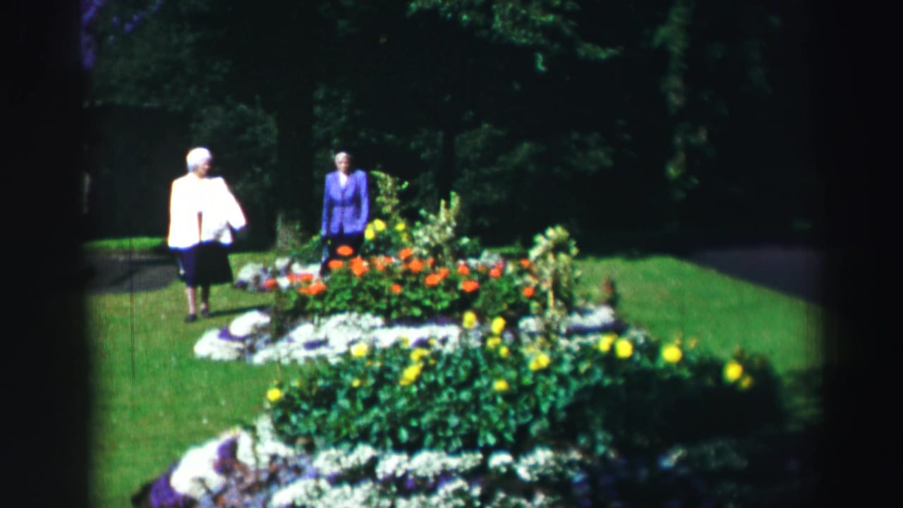 Two women pause by a bright garden bed with orange and yellow flowers.