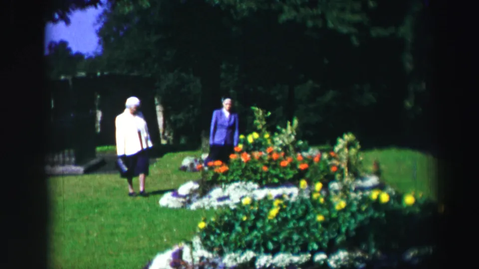 A woman in a light jacket and another in a blue suit walk beside flowers in 1952 Edinburgh home-movie footage.
