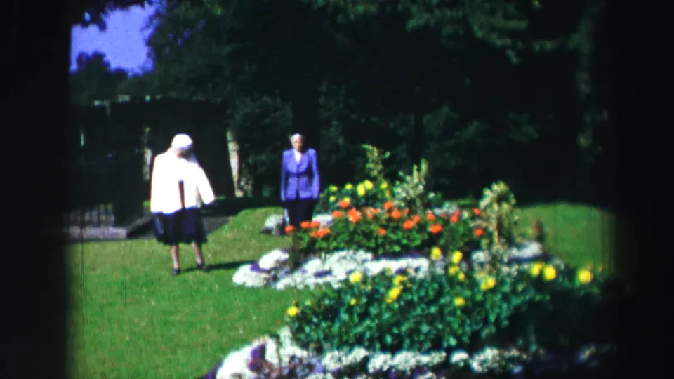 Two women remain near the colorful garden planting, with shaded trees and bright lawn visible behind them.