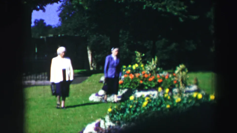 Two elderly women in jackets and dark skirts move through a sunlit garden in supported 1952 Scotland home-movie footage.