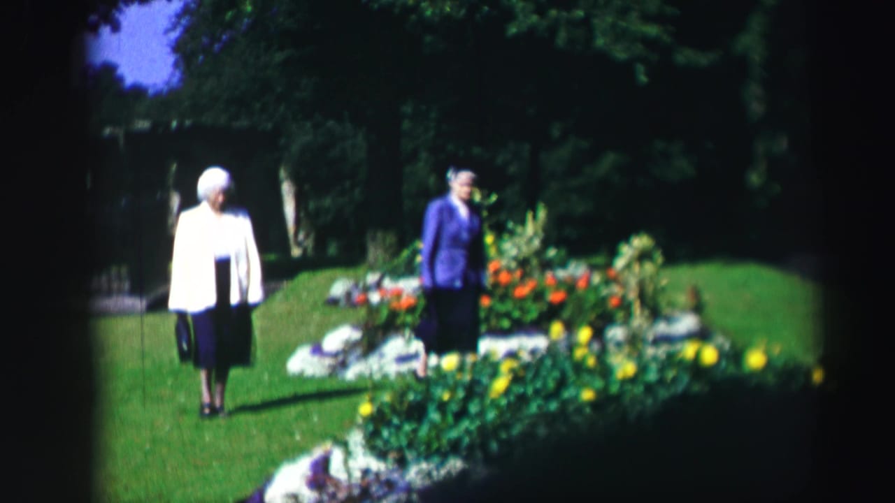 Two elderly women walk beside a bright flowerbed with trees behind them.