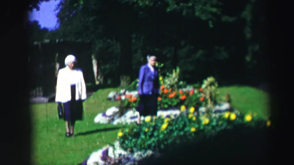 Two elderly women continue across a manicured garden with red and yellow flowers in 1952 Scotland home-movie footage.