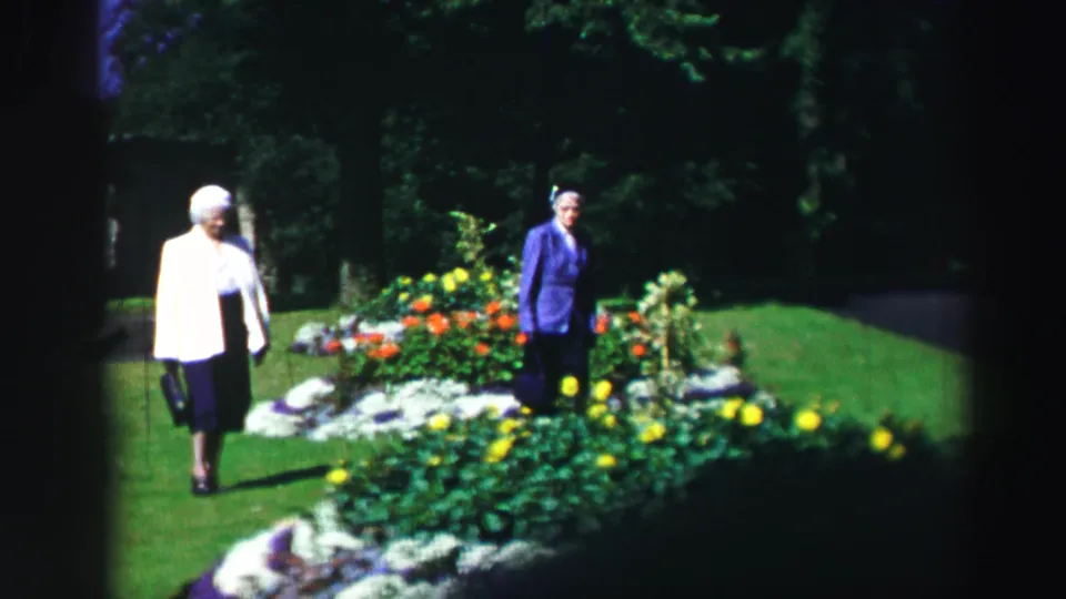 Two elderly women pause near a colorful garden bed, with deep green trees behind them in supported 1952 Scottish footage.