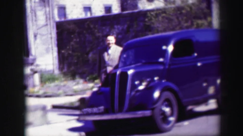 The man pauses again beside the van's front end, with the stone building and small planted strip behind him.