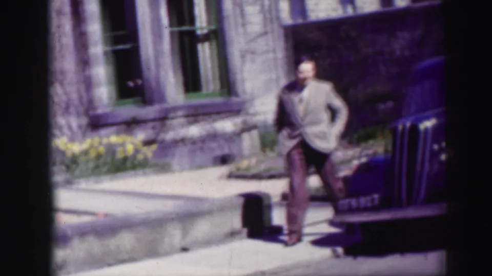 The man steps along the pavement near a green-framed window and yellow flowers, with the blue van partly visible at frame edge.