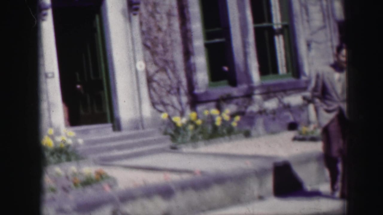 Man in a light suit stands near a stone building and flower beds beside a sidewalk.