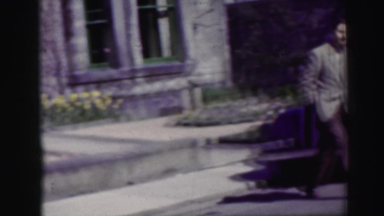 A man in a light suit walks through a 1952 Scotland street scene, partly cropped beside stone facades and greenery.