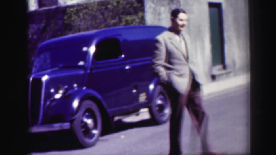 In a 1952 Scotland street view, a suited man walks beside a dark rounded vintage car near stone buildings.