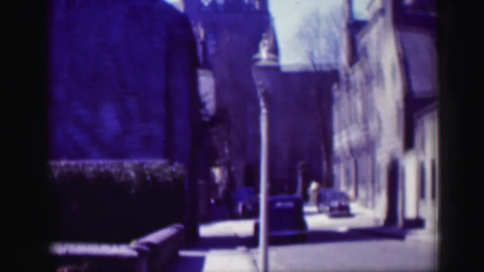 A distant 1952 Scotland street view shows the suited figure near stone buildings, a sidewalk, hedges and a vertical pole.