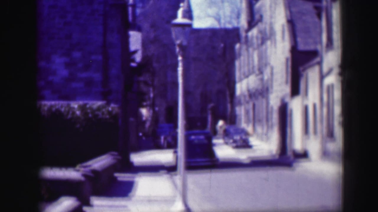 1952 Edinburgh street view with stone buildings, a lamppost, parked cars, and a distant pedestrian in soft 8mm color.