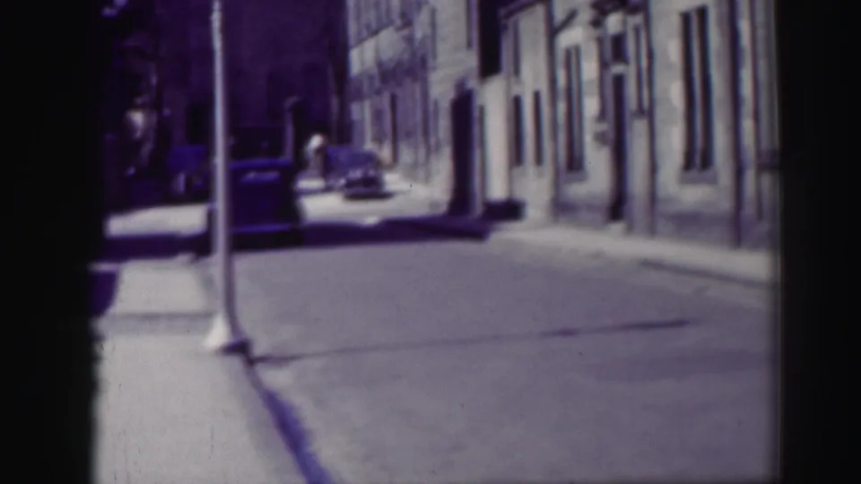 1952 Edinburgh street frame with a tall lamppost in front, parked cars along the curb, and a distant pedestrian near stone buildings.