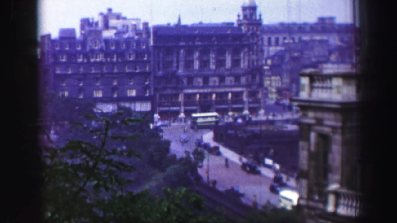 High-angle 1952 Edinburgh street view with stone buildings, vintage cars, a bus, and foliage in the lower frame.