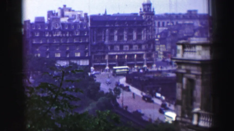 High-angle 1952 Edinburgh street view with stone buildings, vintage cars, a bus, and foliage in the lower frame.