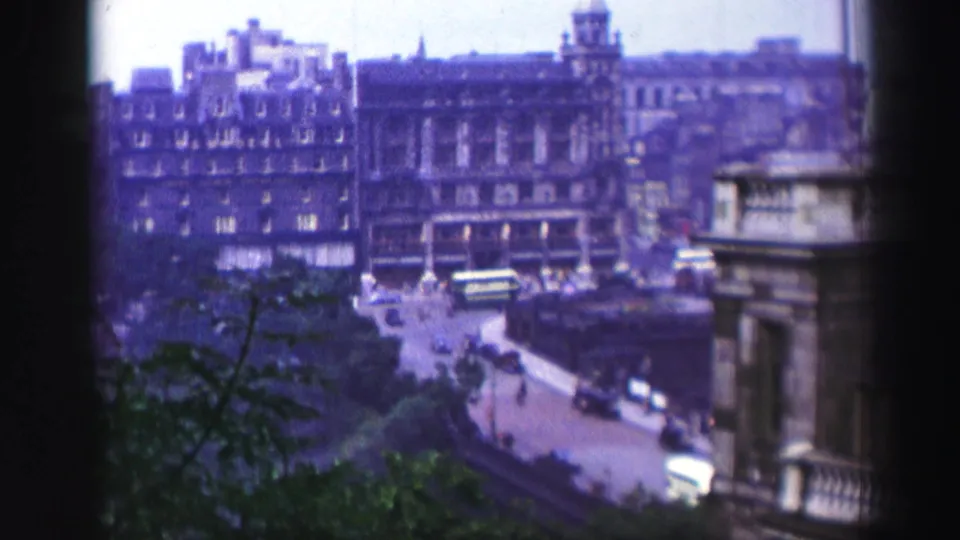 Soft 8mm frame of an Edinburgh street, showing vintage buses, cars, stone buildings, foliage, and a foreground building edge.